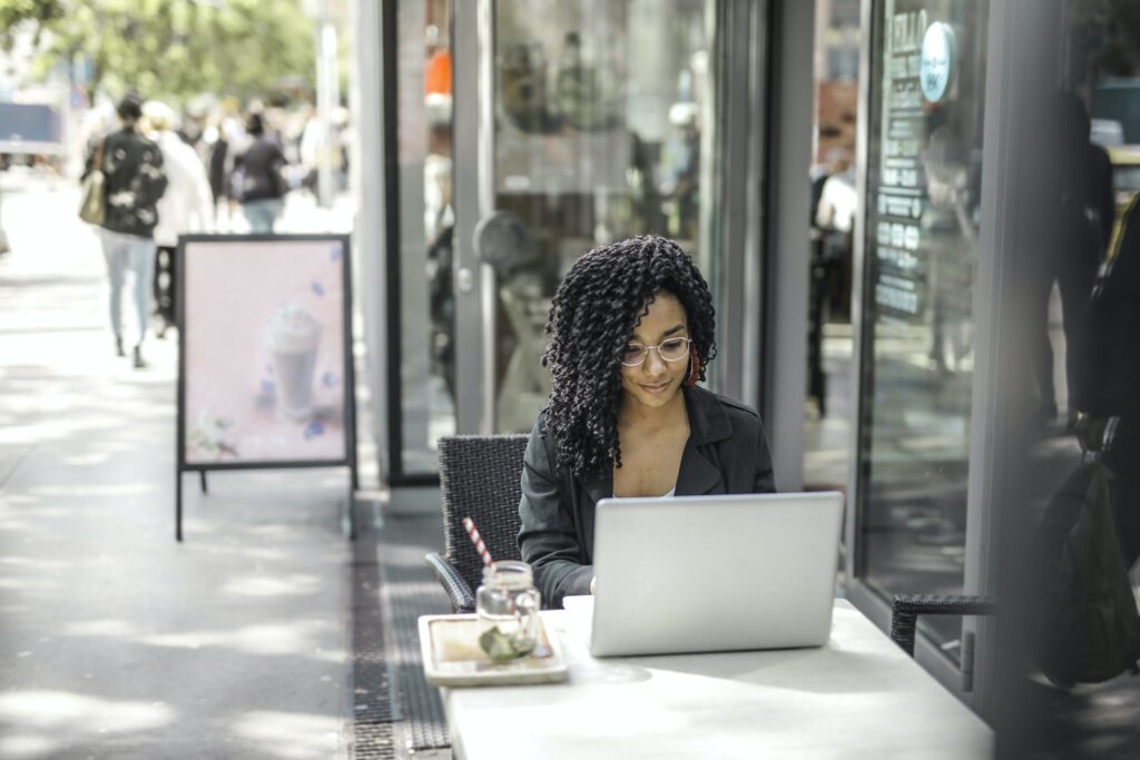 High angle of pensive African American female freelancer in glasses and casual clothes focusing on screen and interacting with netbook while sitting at table with glass of yummy drink on cafe terrace in sunny day